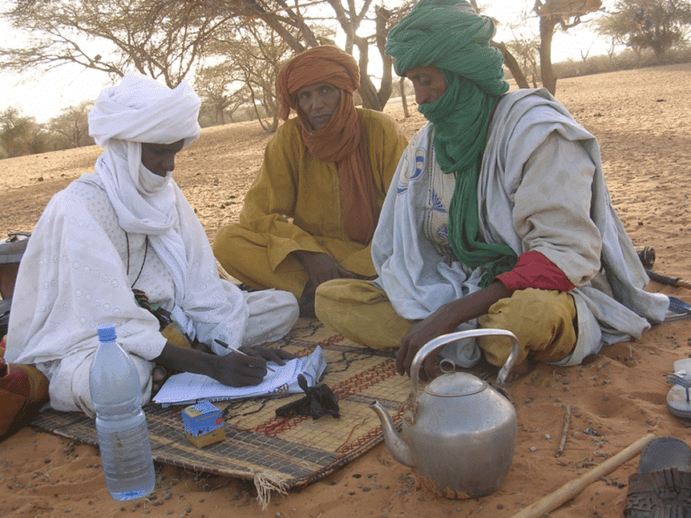 An image of local people being interviewed by a fieldwork taking notes - they are sat cross-legged on the ground