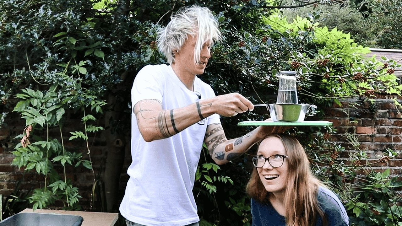 Tom holding a glass of water and a colander over Fiona's head!