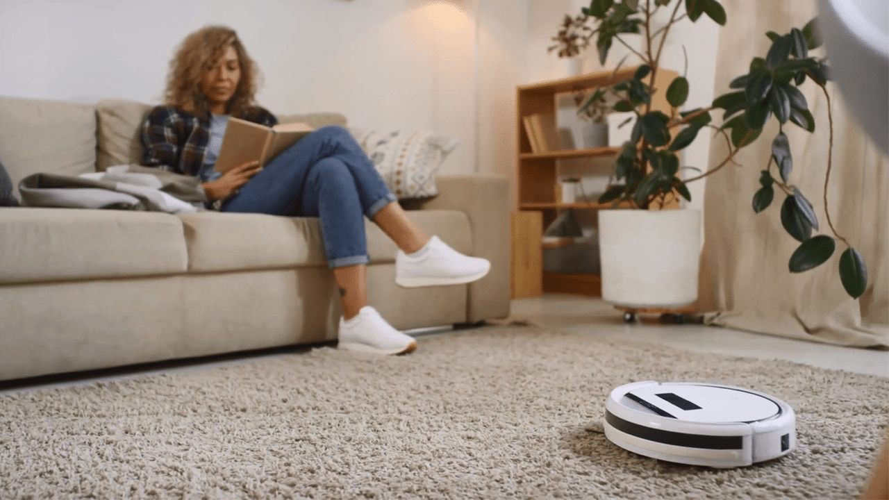 A woman sits on a sofa while a 'robot' vacuum cleaner hoovers the floor. Featured image for the video 'The Future of Automation in the Home'.