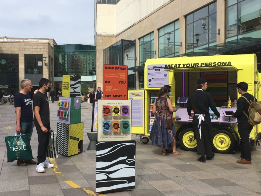 A yellow horsebox surrounded by display boards with information about the health and environmental impacts of meat consumption. Several people are reading the information on the boards. 