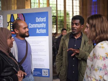 Two men and two women stand together having a conversation next to a poster about the Science Together programme, in a large historic stone hall. 