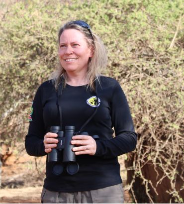 A portrait style photograph of Professor Amy Dickman standing in the African Savannah, holding a pair of binoculars.
