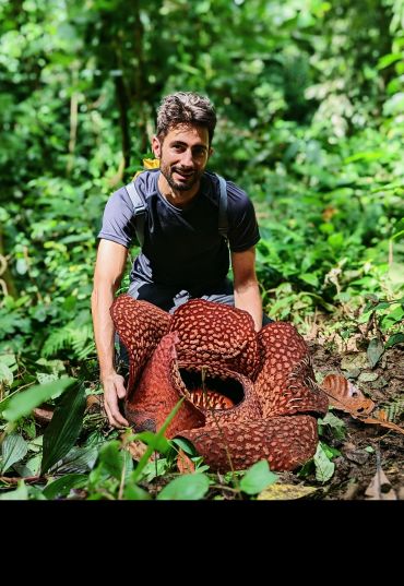 A man stands behind a large five-lobed flower in a jungle. The flower is wider than he is.