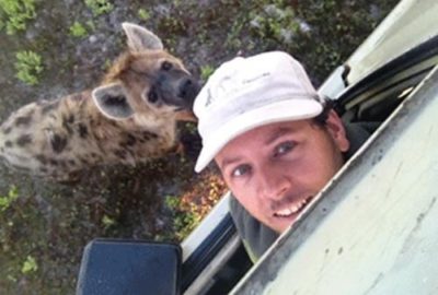 Egil Droge. Man smiling at the camera through a car window, with a hyena in the background.