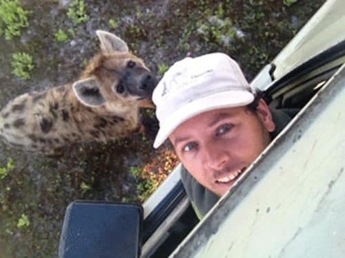 Egil Droge. Man smiling at the camera through a car window, with a hyena in the background.