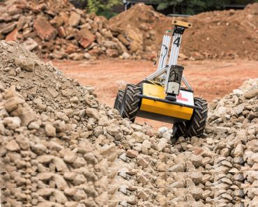 A small box-like robot with four tyres drives over piles of gravel and rubble.