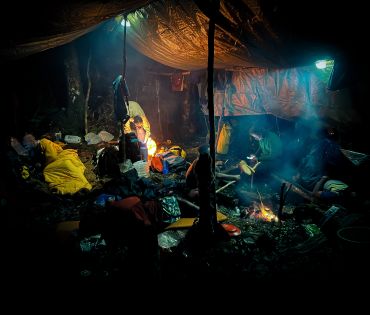 The interior of an open-sided tent in the middle of a jungle. Members of the expedition are visible sitting or in sleeping bags. 