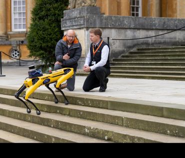 A dog-like robot with a rectangular body and four jointed legs stands on the steps leading up to a stately home. It faces two men who are crouched down in front of it.