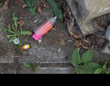 A broken neon colored electronic cigarette vape has been discarded in dirt and weeds. The battery from the device has fallen out too.