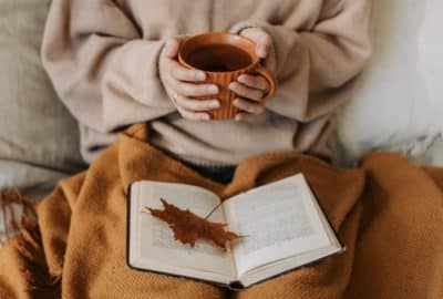 Woman holding a coffee up with a blanket and open book on lap