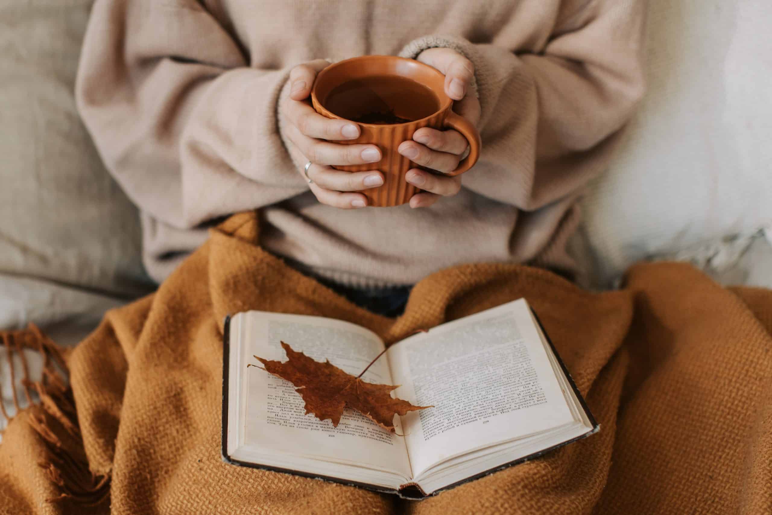 Woman holding a coffee up with a blanket and open book on lap