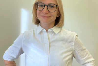 A woman with short blonde hair and wearing glasses is smiling, with her hands on her hips.