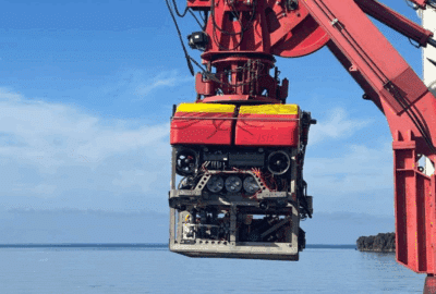 A Remotely Operated Vehicle (ROV) is suspended from the side of a research ship on a sunny day in the Aegean Sea, just off the coast of Santorini.