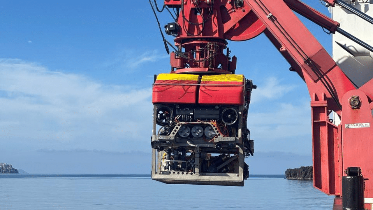 A Remotely Operated Vehicle (ROV) is suspended from the side of a research ship on a sunny day in the Aegean Sea, just off the coast of Santorini.