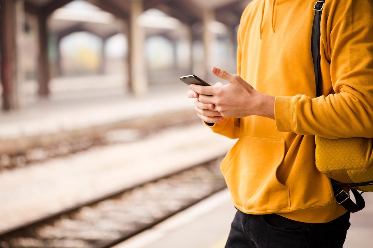 A man dressed in a yellow jumper stands on a train station platform holding a mobile phone in his hands.