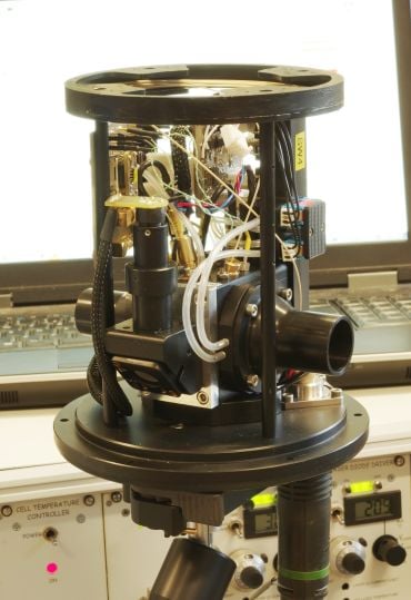 A close-up view of a molecular flow sensor, showing its exposed wiring, tubing, and optical components, mounted in a cylindrical frame with control equipment in the background.