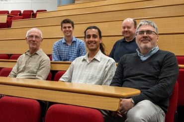 Five people seated in a lecture theatre with red chairs and wooden desks, arranged in two rows and smiling towards the camera.