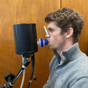 A volunteer wearing a nose clip breathes into a black cylindrical device connected to tubes and sensors, as part of a computed cardiopulmonography (CCP) breath test.