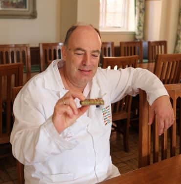 Professor Charles Spence, a white man wearing a white lab coat. He sits in a dining chair at a long table and holds a bar of Dubai Chocolate.