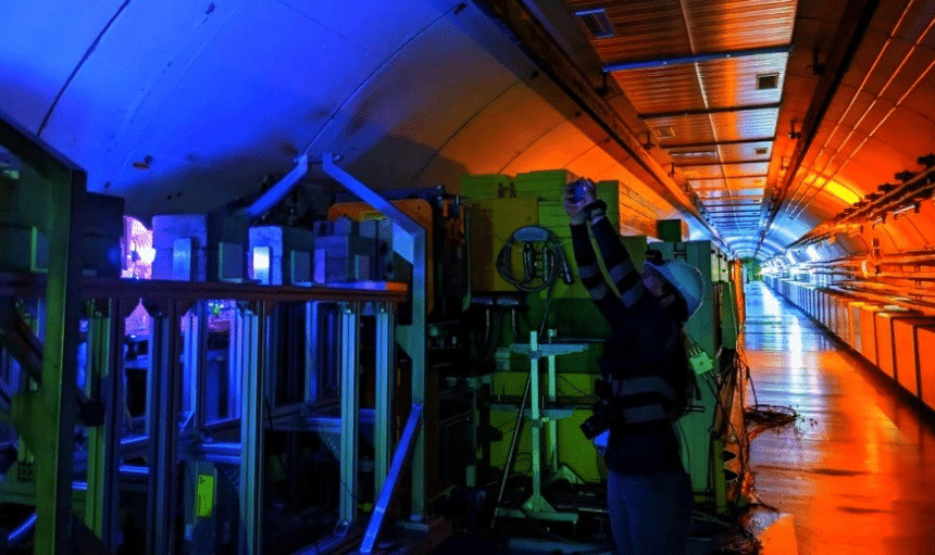 A researcher wearing a hard hat stands next to scientific machinery in a tunnel illuminated by orange and blue lighting. 