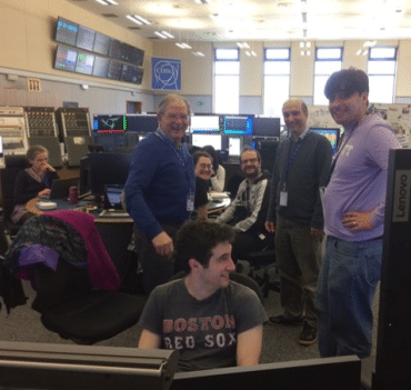 A group of male and female researchers sit and stand around a bank of computers in the CERN control room.
