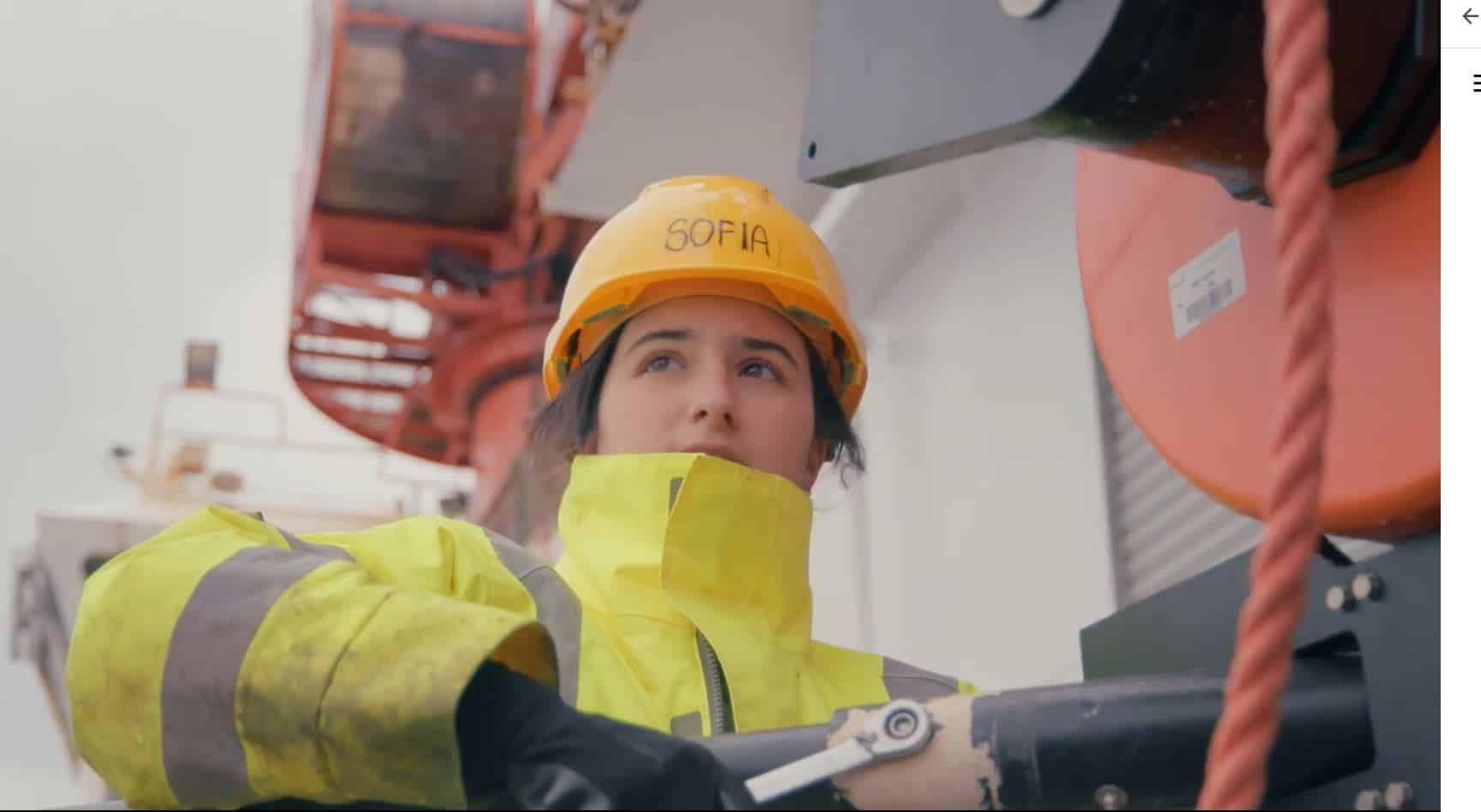 Sofia della Sala aboard a research vessel just off the coast of Santorini, wearing an orange hardhat labelled 'Sofia' and a yellow high visibility coat.