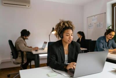 Four colleagues working in an office on their laptops. The main figure is a smartly-dressed young woman wearing headphones and typing on her laptop.